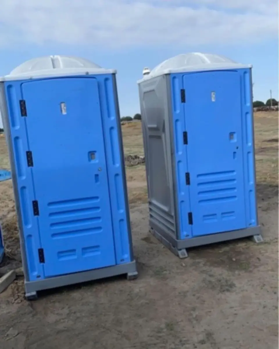 Two standard porta potties standing on an open dirt lot