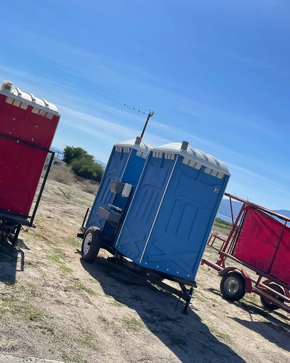 Blue porta potty mounted on a small towable trailer on a dirt lot